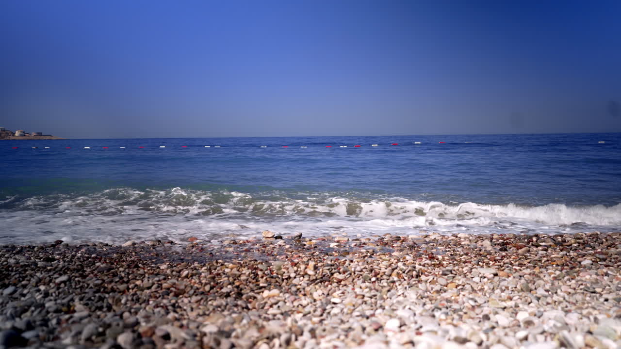 아드리아해의 파도는 돌과 자갈이 있는 바위 해변에 부딪혀 하얀 거품으로 씻겨지고, 해안 가까이에 떠 있는 빨간색과 흰색 부표 줄, 위의 구름 없는 푸른 하늘, 정적 4k