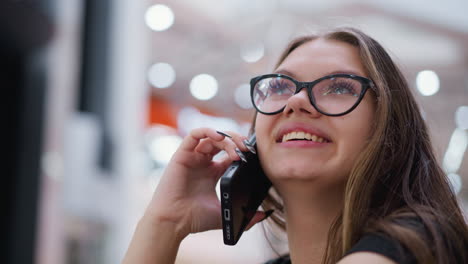 primer plano de una mujer con camiseta negra y gafas hablando por teléfono, riendo, con enfoque en el fondo y telón de fondo borroso con efecto de luz bokeh y ascensor en movimiento