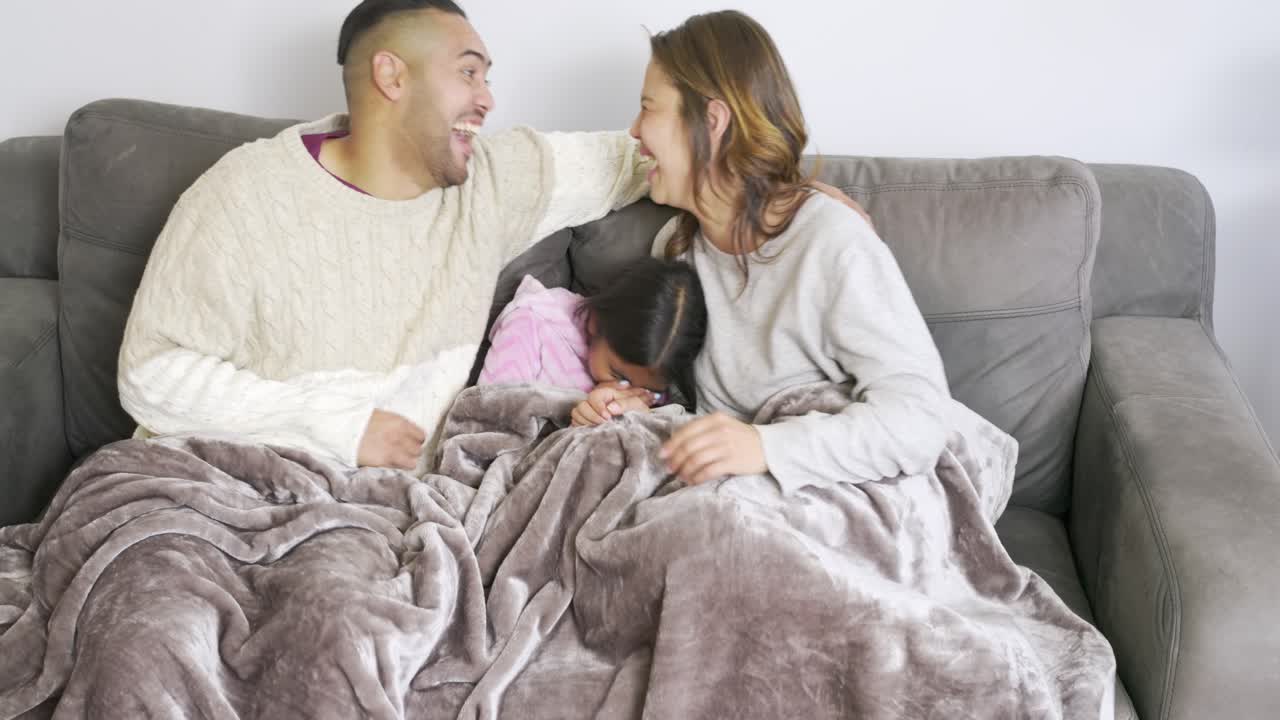 Una famiglia eterogenea si sistema sotto una coperta sul divano, premendo il telecomando e ridendo per uno spettacolo fuori campo.