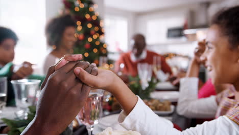 Selektives-Fokusdetail-Von-Mutter-Und-Tochter,-Die-Händchen-Halten,-Während-Ihre-Familie-Aus-Mehreren-Generationen-Am-Weihnachtstisch-Das-Tischgebet-Betet,-Aus-Nächster-Nähe