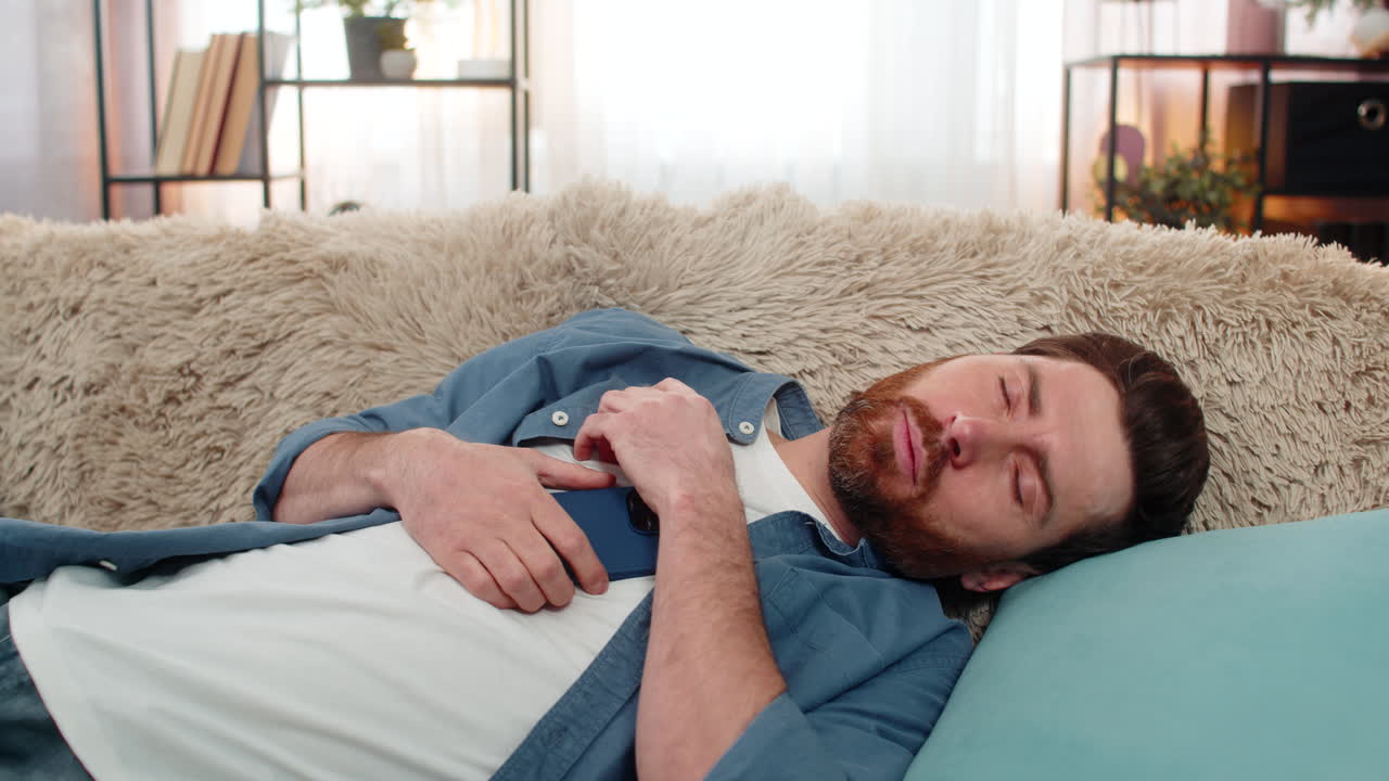 Young man unexpectedly falling asleep on sofa while holding smartphone in hand tired and exhausted