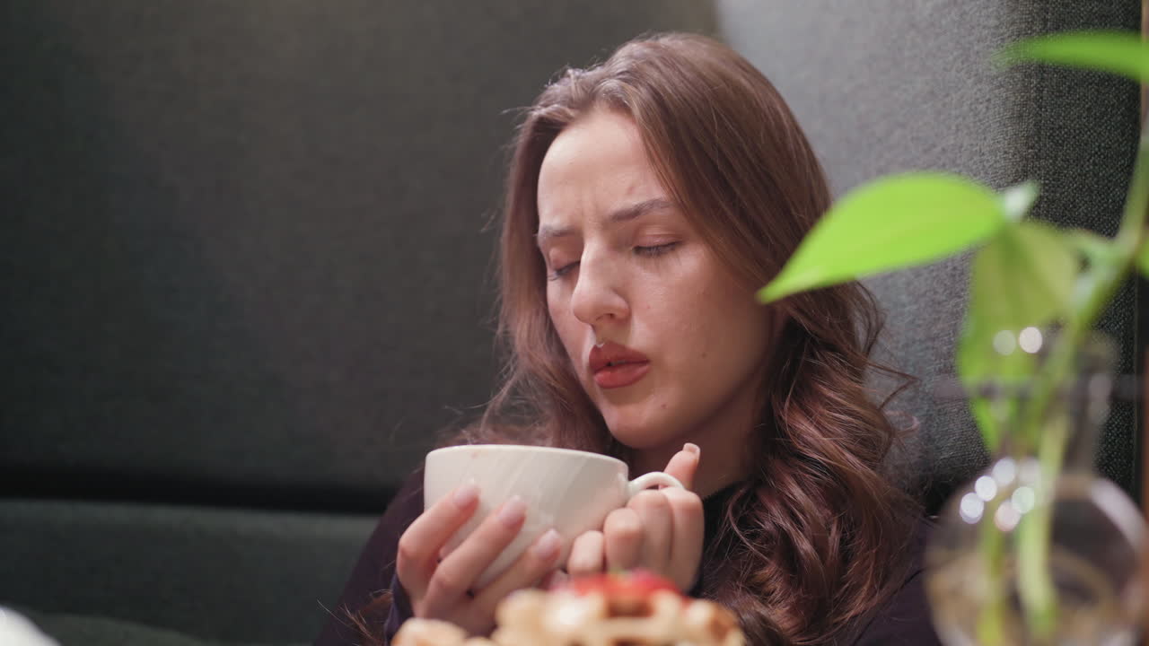 Lady with long brown hair holds white cup of hot latte and gently blows on it to cool down while seated in cozy booth, with glass vase containing green plant on table and waffles in foreground
