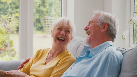 Una-Pareja-De-Ancianos-Jubilados-Sentados-En-Un-Sofá-En-Casa-Viendo-Televisión-Y-Riendo-Juntos