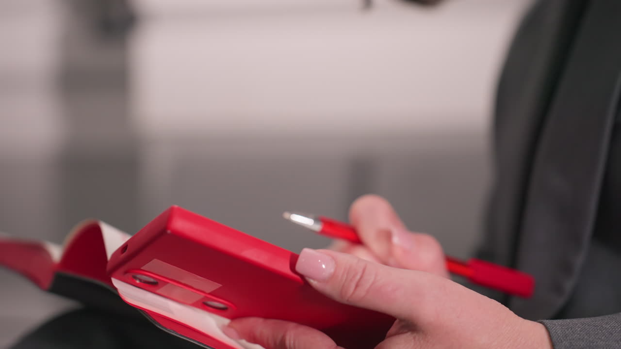 Close-up of person holding red phone with one hand and using red pen to write in notebook with other hand. Focus on professional work, ideal for office tasks, meetings, and brainstorming sessions