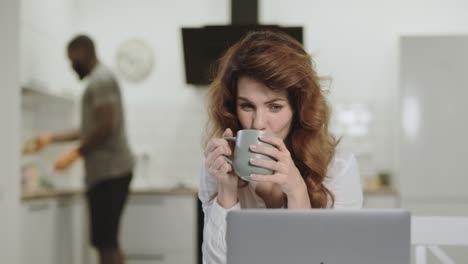 mujer blanca sonriente leyendo noticias felices en la computadora portátil en la cocina de la mañana.