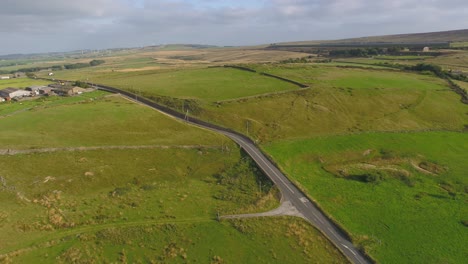 Drone-footage-descending-and-panning-showing-the-West-Yorkshire-countryside-in-England,-UK-including-a-farm,-moorland,-country-road-with-moving-cars,-dry-stone-walls,-telegraph-poles-and-wind-turbines