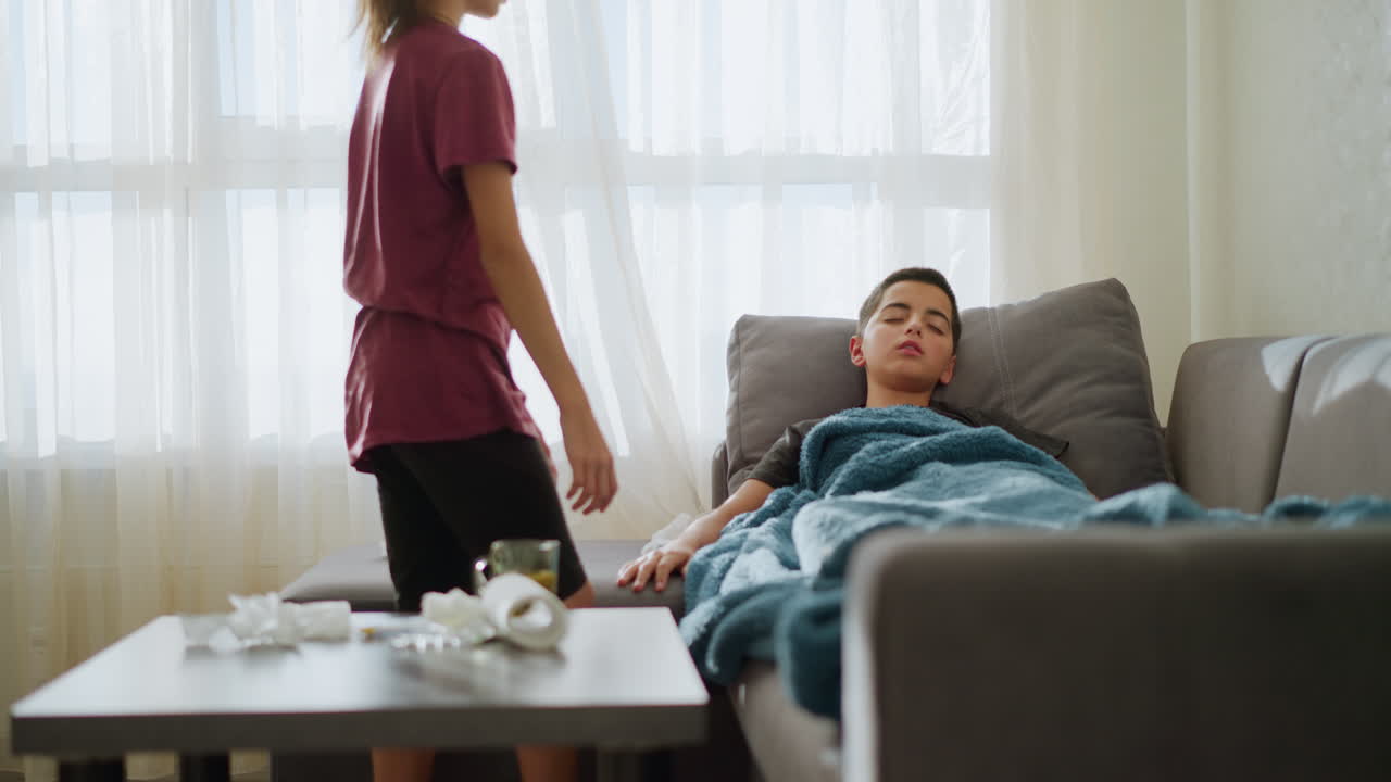 Little cousin walking toward her brother who is sleeping on the couch, she adjusts the blanket while used tissues are scattered around, showing a caring moment