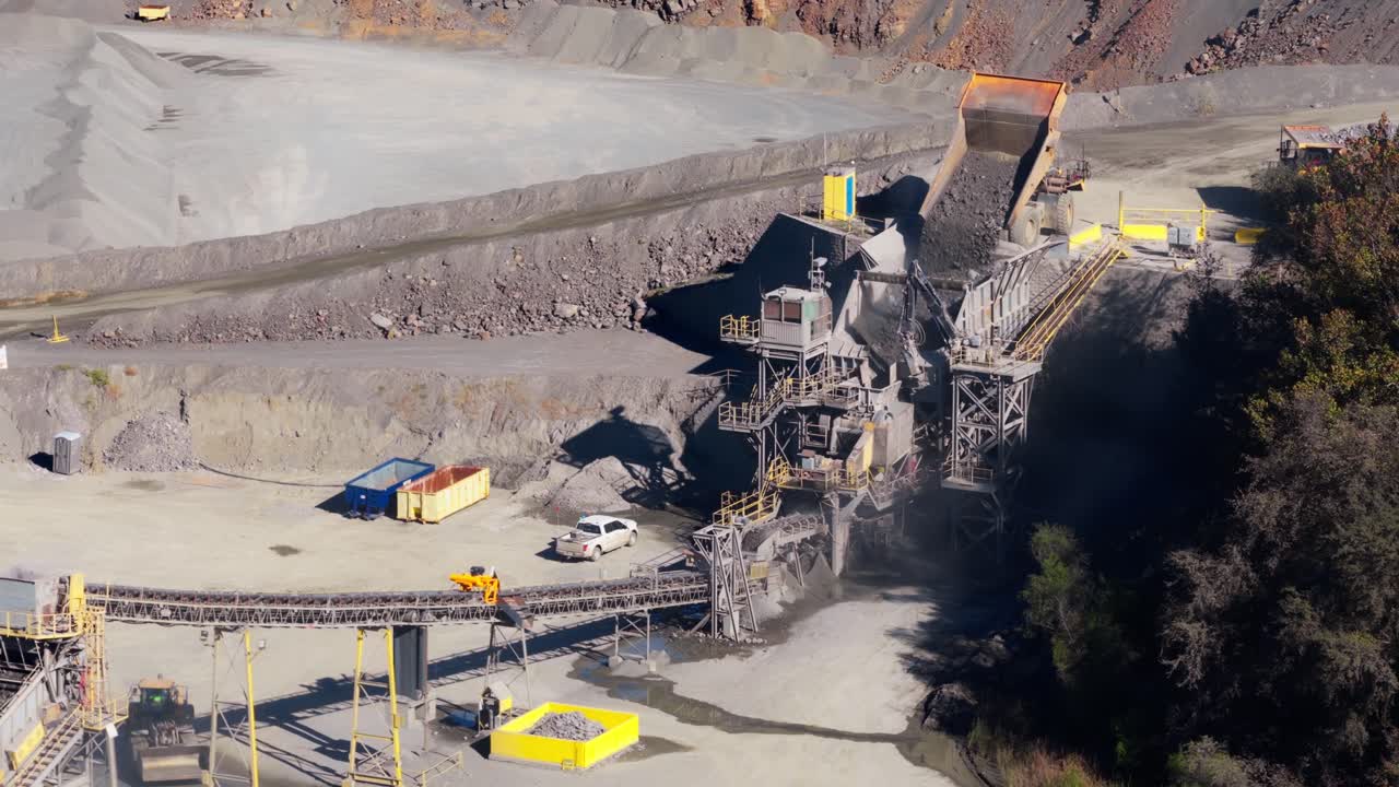 Wheel loader moving gravel beside conveyor systems at quarry as large dump truck emptys fill into hopper
