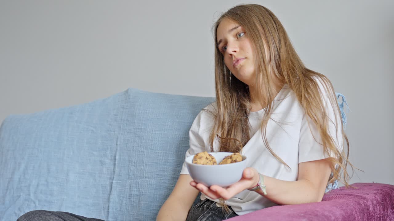 Donna attraente a casa che usa il telecomando della televisione mentre tiene in mano una ciotola di biscotti.