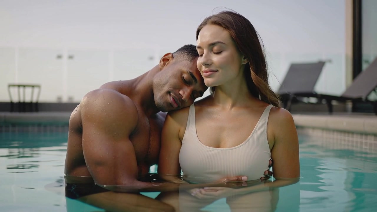 Leaning man resting head on her shoulder, both standing in rooftop pool, relaxing in swimwear