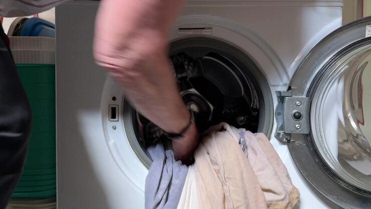 Close view of a woman's hands opening a front-loading washing machine and placing bed linen inside, showing an everyday laundry and household routine. Static shot.