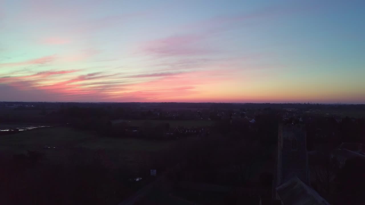 Drone shot of beautiful pink sky tracking down to show rural English village with big church and graveyard