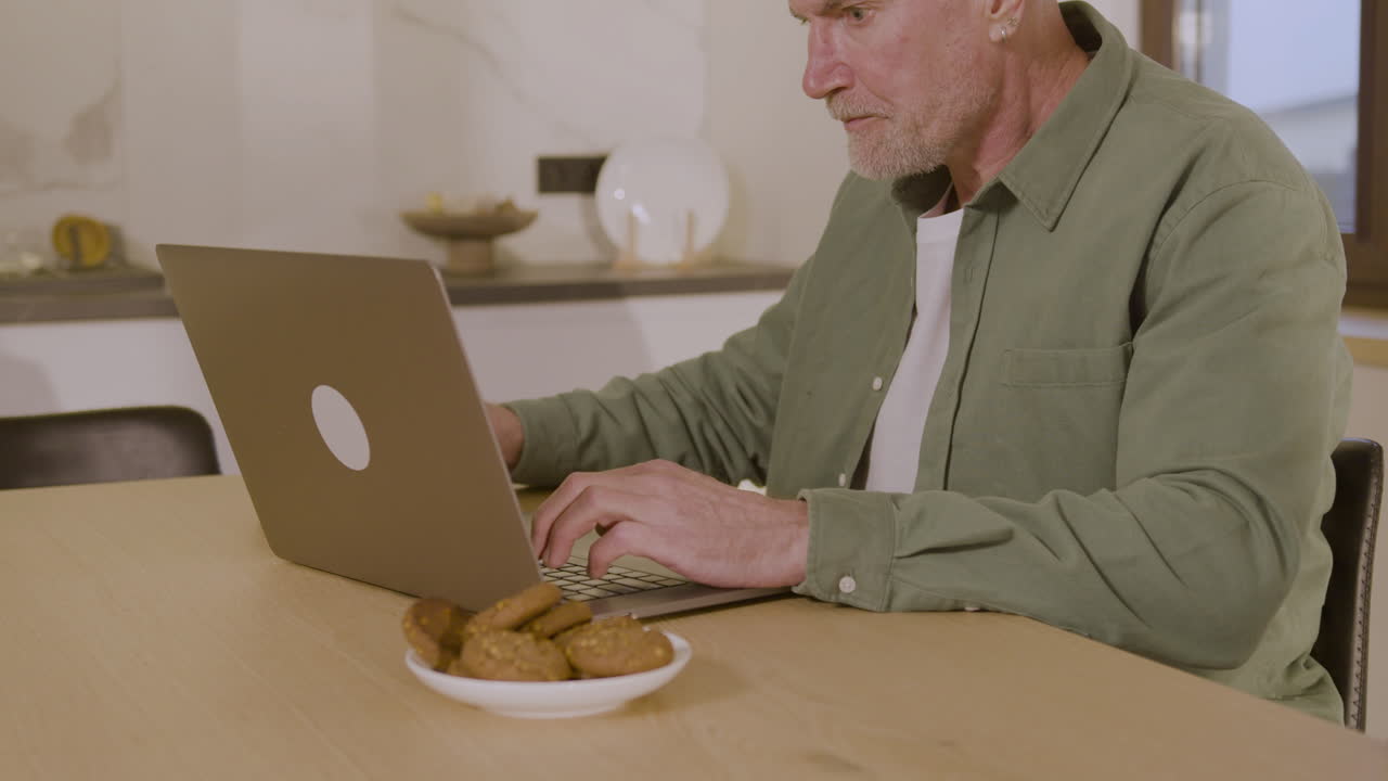 Happy Elderly Man Sitting On Chair In Kitchen, Using Laptop Computer And Drinking Tea 1
