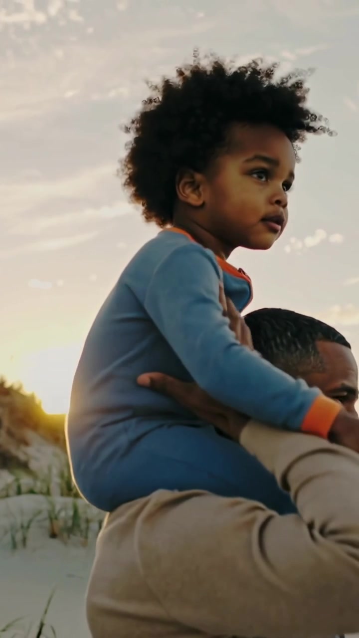 Vídeo vertical: Pai sorridente levantando criança com roupa azul nos ombros em duna ao pôr do sol para ver a costa.