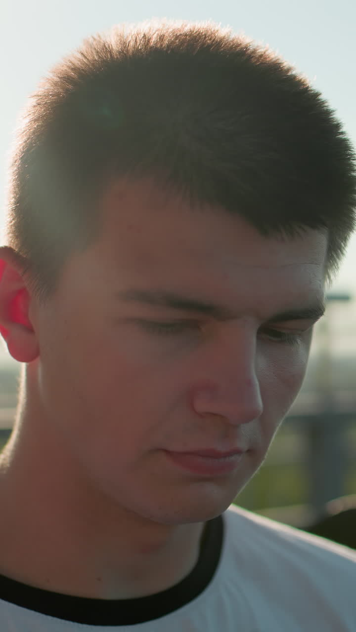 primer plano de un hombre con camiseta blanca mirando hacia abajo con la luz del sol reflejándose suavemente en su cara, mostrando una expresión tranquila e introspectiva, con fondo con vegetación y barandilla de hierro
