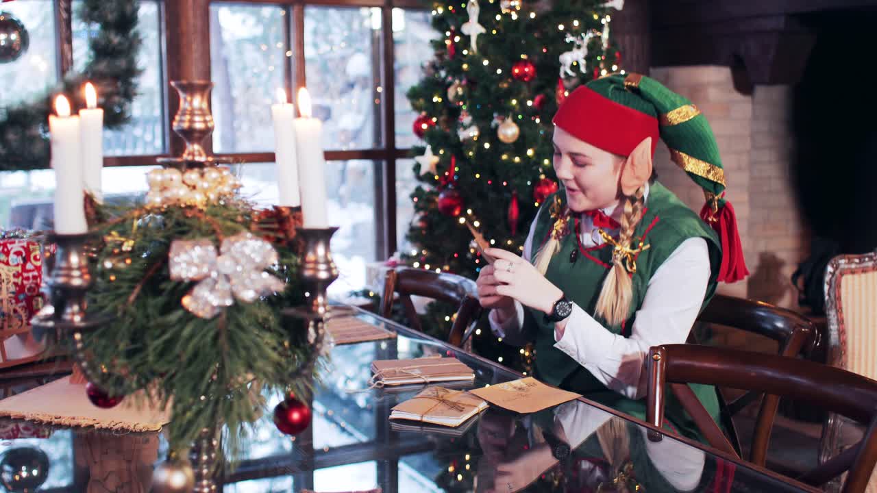An elf girl sits in a room with a Christmas tree and looks through the letters to the children of Santa Claus