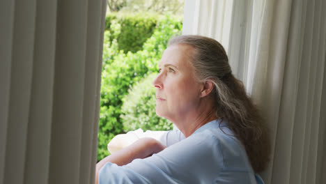 Senior-caucasian-woman-wearing-blue-shirt-and-looking-through-window-in-living-room