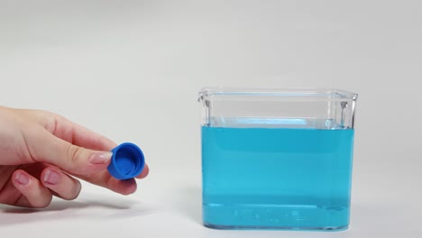 a hand places a blue plastic cap in a water-filled container, demonstrating buoyancy principles under bright lighting