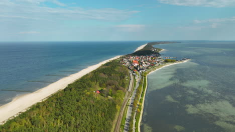 Aerial-view-of-a-narrow-strip-of-land-with-a-road-dividing-the-dense-greenery-and-a-coastal-village-from-the-expansive-sandy-beach-and-open-sea-on-one-side-and-the-calm-bay-on-the-other---Kuźnica,-Hel