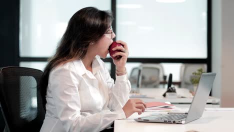 a beautiful and healthy businesswoman taking a bite of an apple while working on a laptop business assignment.