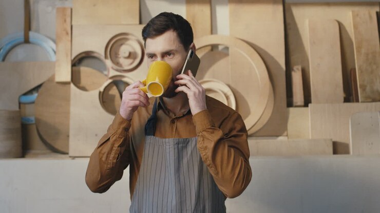 Portrait of caucasian bearded man in apron talking on the phone and drinking coffee in carpentry workshop
