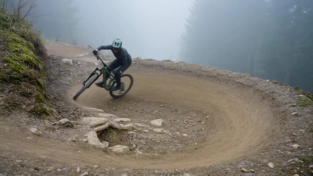 Avvicinandosi a una sponda su un sentiero nebbioso, ciclista MTB con casco verde che si inclina, spostandosi sulle radici mantenendo la presa