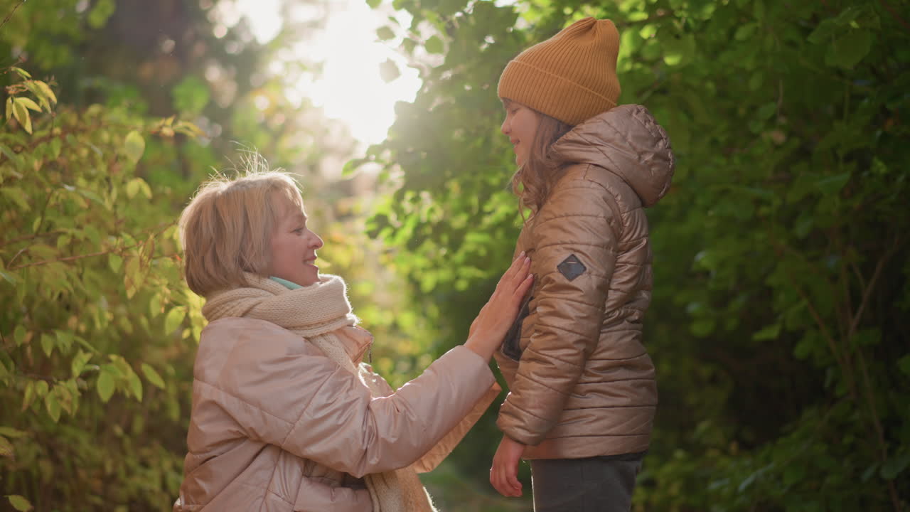 lovely moment with mother kneeling in autumn woods as daughter turns to kiss her cheek, warm sunlight filtering through colourful foliage, expressions tender and joyful in natural setting