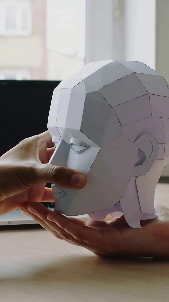 Vertical video: Bringing paper lowpoly head down, hands balancing model on palm at desk, laptop