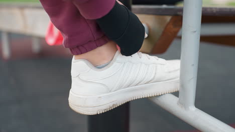high angle closeup of female athlete tying sneaker lace on iron exercise bar rail in urban outdoor fitness park before starting strength training session under open sky near modern apartment complex