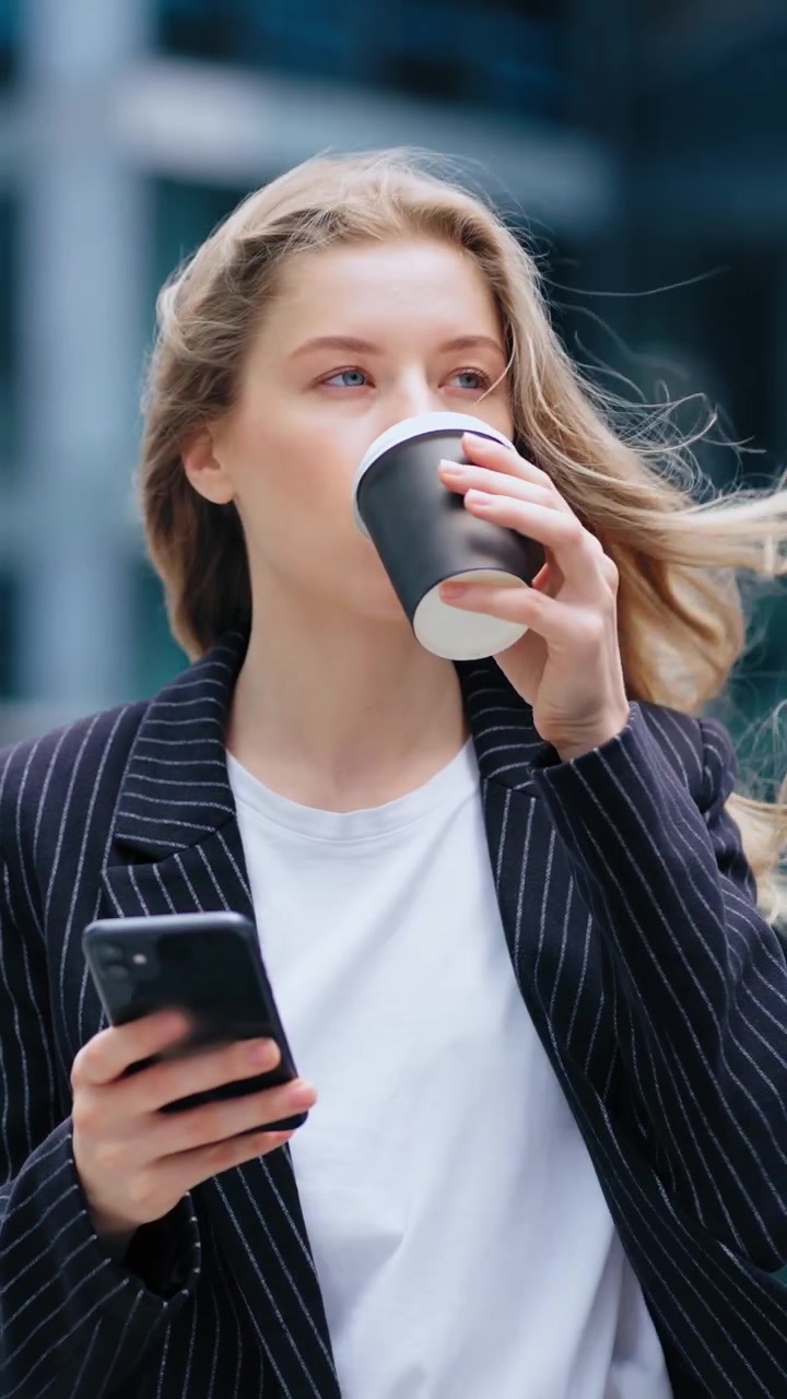 Ung forretningskvinde bruger smartphone og drikker kaffe udendørs.