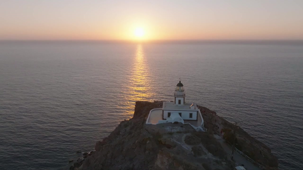 Riprese con drone del sole che tramonta nel Mar Egeo, rivelando il faro di Santorini e le spettacolari scogliere della caldera avvolte nella luce dorata del tramonto.