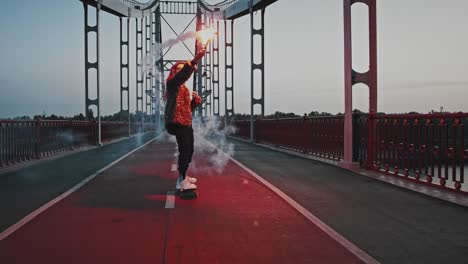 young hipster woman in informal clothes is skateboarding on bridge while holding glowing red signal flare and waving it