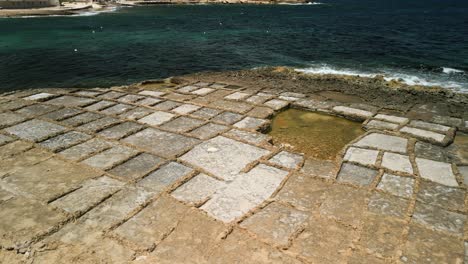 Rotating-aerial-footage-of-rectangular-salt-pans-with-sea-waves-on-the-coast-of-Gozo,-Malta