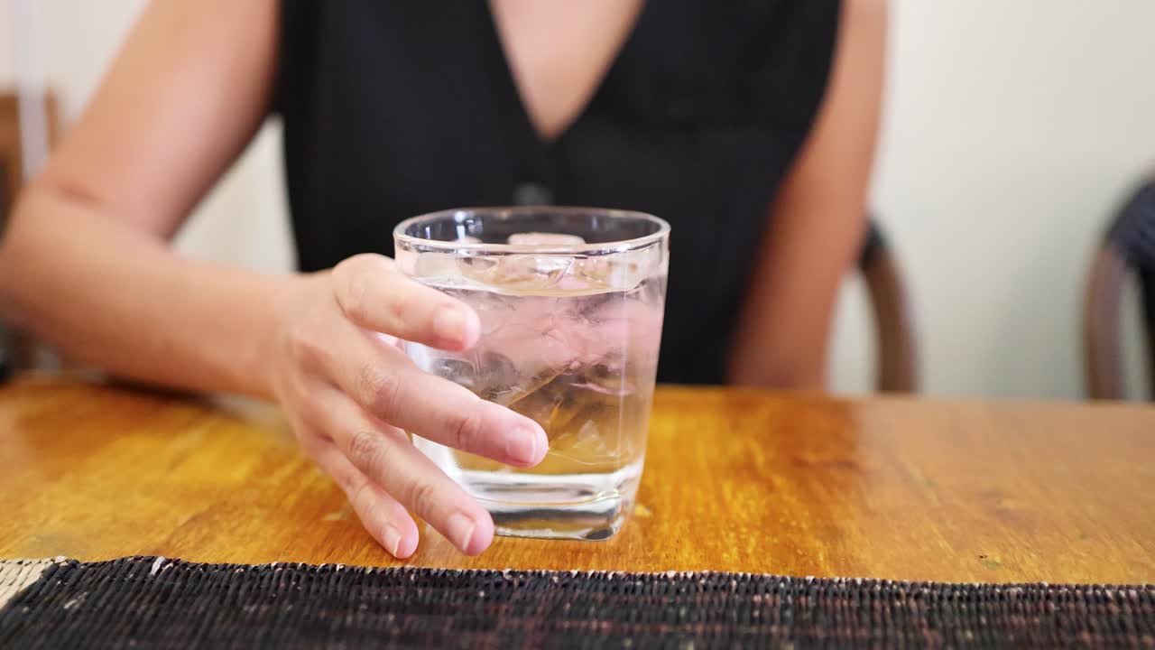 A woman in a sleeveless black top reaches for and lifts a glass of ice water from a wooden table in a well-lit indoor setting