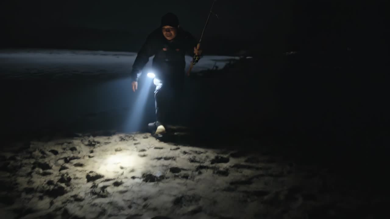 Pescador con abrigo acercándose a la cámara, barriendo la luz, levantando la caña de pescar en una playa nocturna con un señuelo verde.