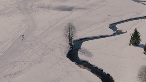 drone orbit shot of a man cross-country skiing with his dog in a snowy landscape with a river