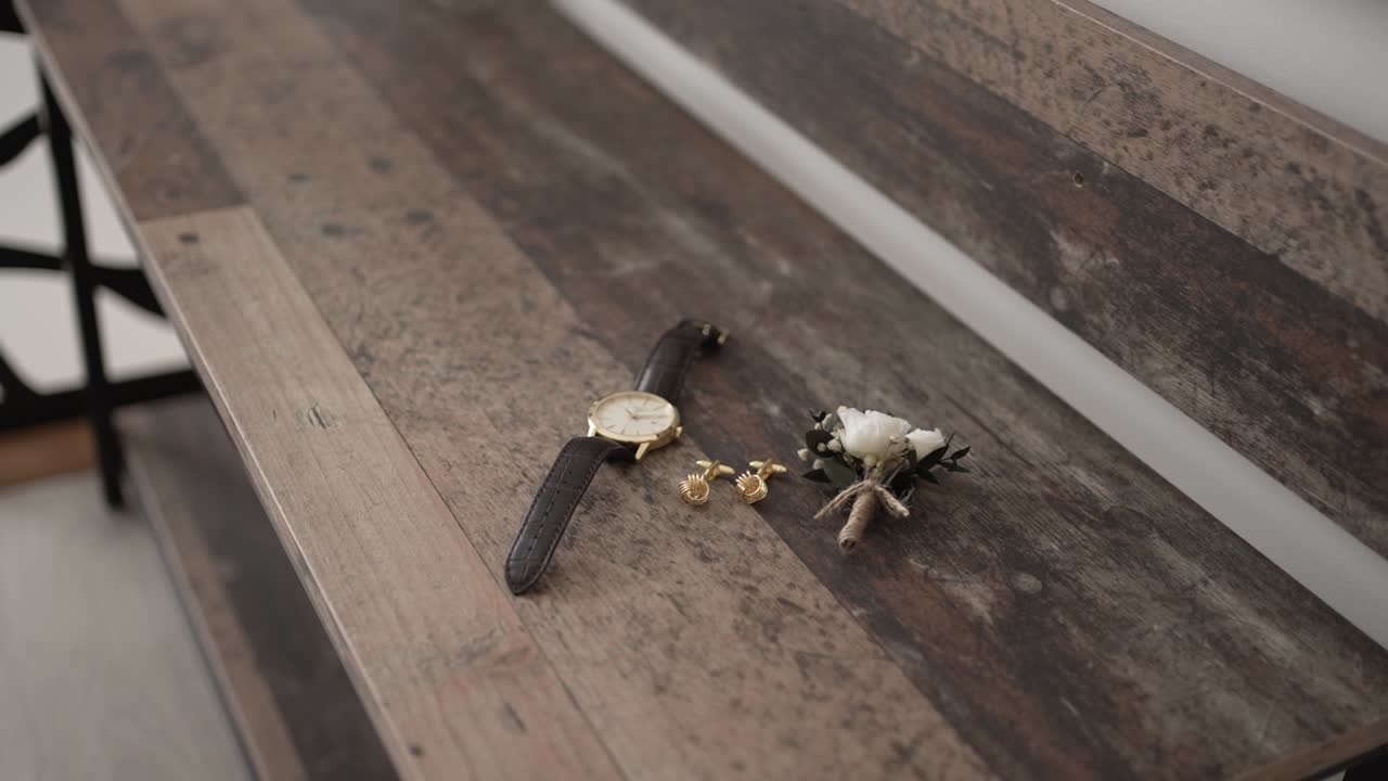 Brown leather watch, gold cufflinks, and white boutonniere arranged on rustic wood surface