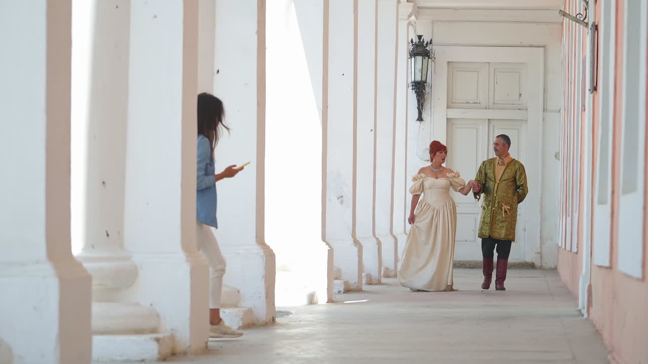 Adult couple in old-fashioned clothes walking by the long hall holding by hands. Girl in modern look appears taking selfie. Present meets past.