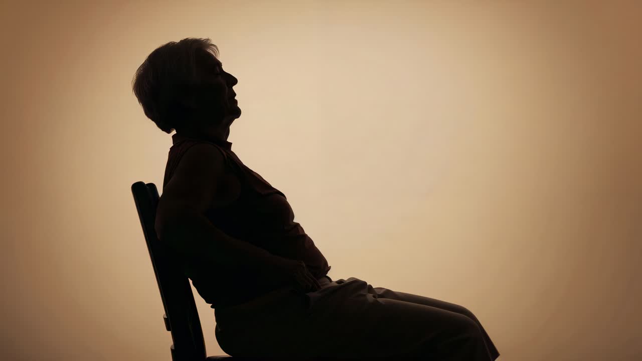Shifting weight senior woman reclining in chair in studio, silhouetted on warm backdrop, copy space