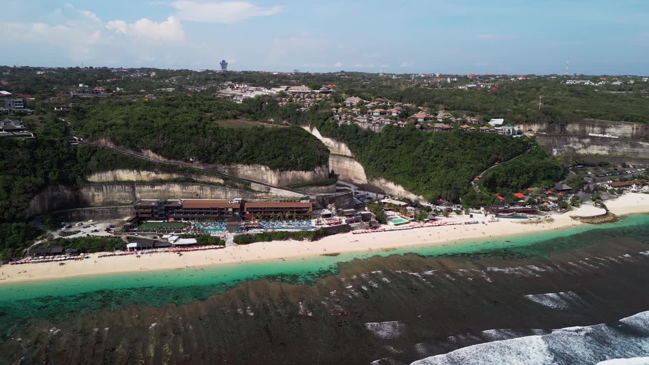 Drone view of Uluwatu's Melasti Beach showing steep limestone cliffs, bright sand, and modern beach clubs with vibrant turquoise waves and panoramic island views