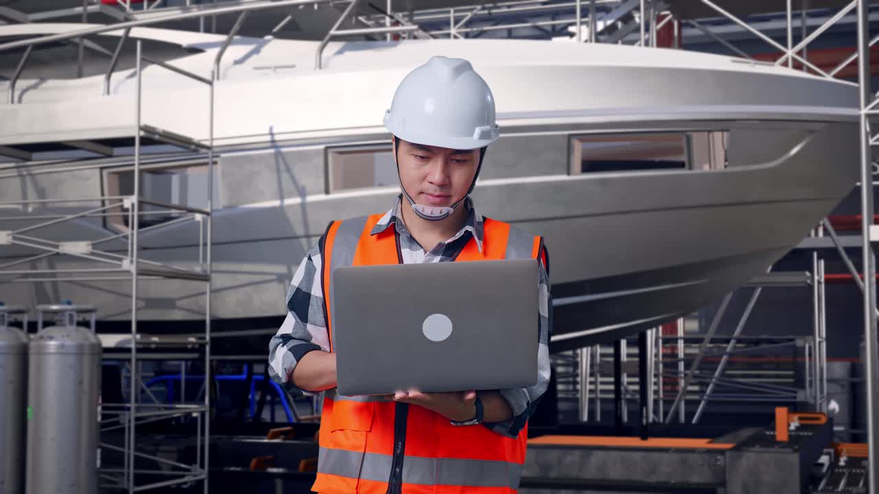 Ingeniero asiático con casco de seguridad trabajando en un portátil de pie con la construcción de un yate en un astillero moderno