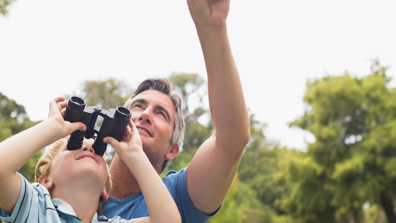 Caucasian father pointing to sky with son looking through binoculars, birdwatching in countryside