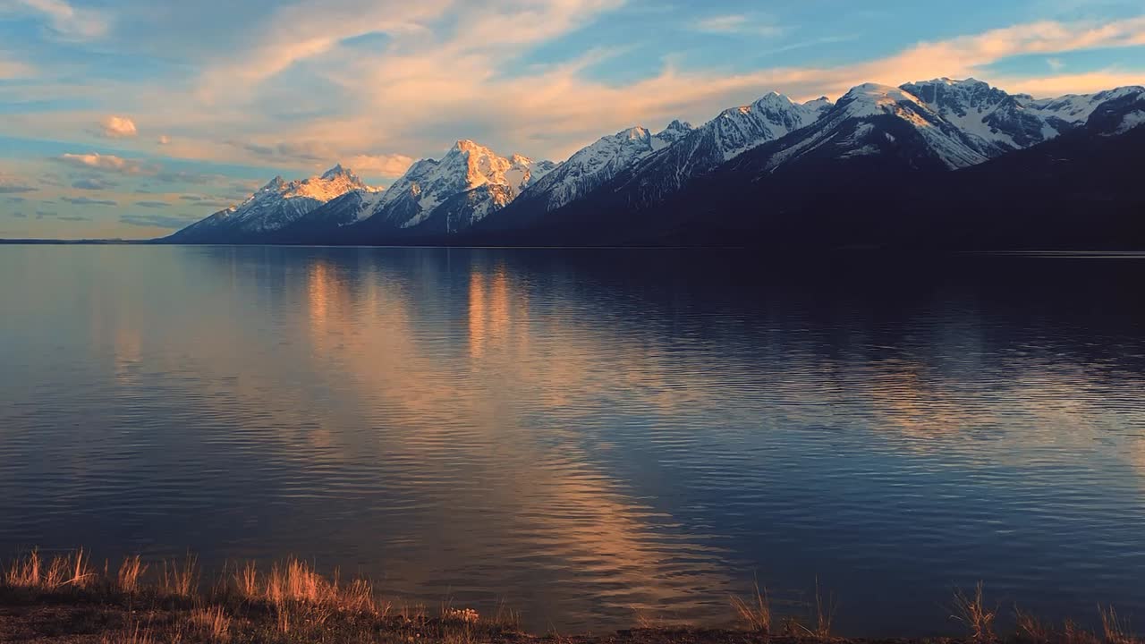 Teton Mountain Range, Wyoming, USA reflected on the surface of a tranquil lake with gently rippling water during sunset. Ideal for nature documentaries, travel content, or background.