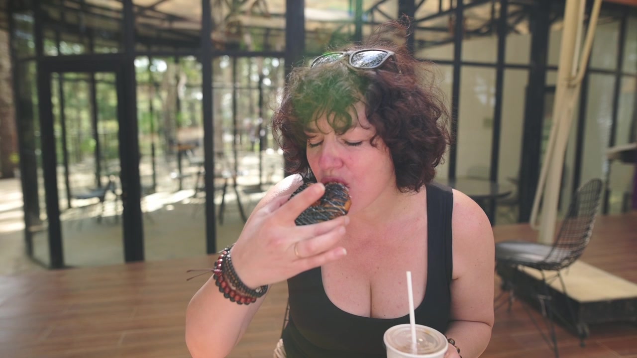 Donna che addenta un grande pezzo di cioccolato con le dita, un'indulgenza giocosa e disordinata catturata in un caffè serra illuminato dal sole. Briciole e glassa sulla mano, una tazza di bevanda sul tavolo e un momento di gioia spontanea.