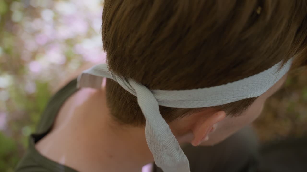 close view of the Tourist loosening headband while sitting on grass alone in the forest and reflecting in nature, preparing for next adventure, with clear sunlight filtering through surrounding trees