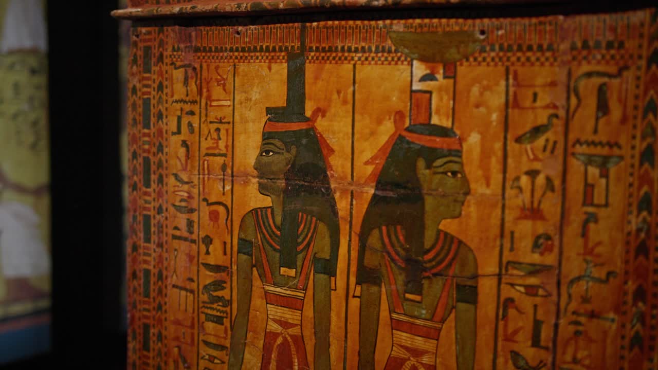 Lähikuva muinaisesta egyptiläisestä puisesta arkun paneelista, jossa on tyylitelty naishahmo perinteisessä profiilissa, koristeltuna päähineellä ja koruilla, monimutkaisten hieroglyfimerkintöjen ympäröimänä.