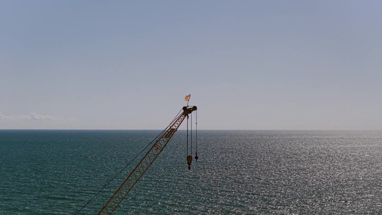 Stationary lattice boom crane with two hanging hooks rises above a sunlit open sea under a clear sky.