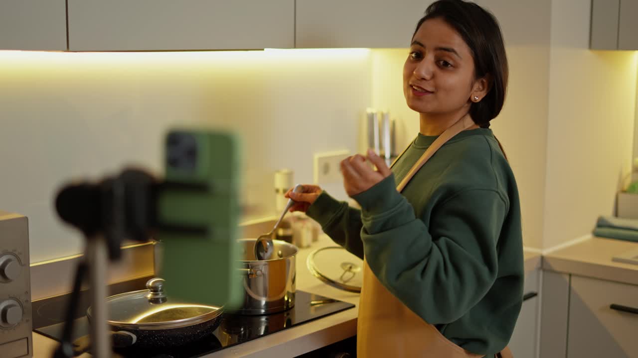 A happy brunette girl with Brown skin in a green jacket and a brown apron broadcasts and broadcasts her culinary blog while cooking in a gray shiny pan in a modern kitchen