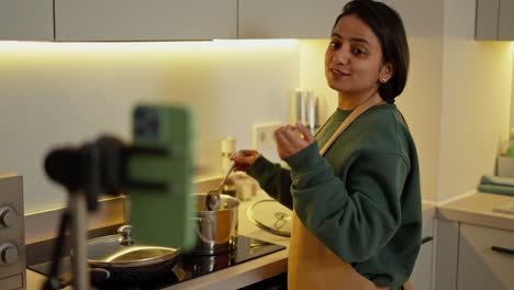 A-happy-brunette-girl-with-Brown-skin-in-a-green-jacket-and-a-brown-apron-broadcasts-and-broadcasts-her-culinary-blog-while-cooking-in-a-gray-shiny-pan-in-a-modern-kitchen