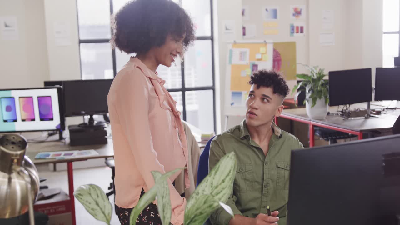 Two happy diverse creative designers in discussion at computer in casual office, slow motion
